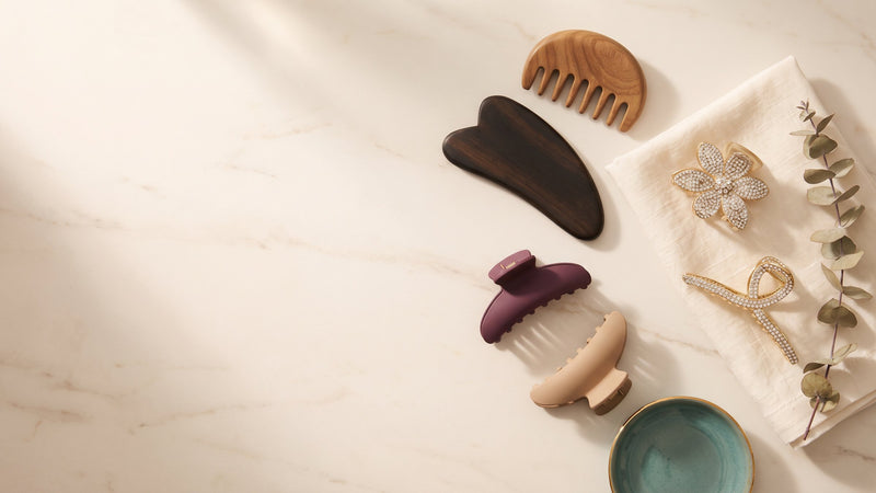 Flat lay of a wooden gua sha board, sandalwood comb, claw clips and a rhinestone flower clip on cream marble